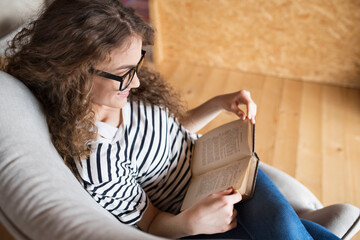 Wall Mural - Female student sitting in armchair, reading book, studying for final exam at university. Beautiful woman relaxing at home.