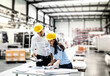 © Halfpoint - Two project managers standing in modern industrial factory, looking blueprints and documents on table. Manufacturing facility with robotics, robotic arms and automation. Storing products and materials