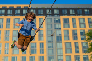 Wall Mural - Kid swinging on chain swing on city kids playground. Swing ride. Cute child having fun on a swing on summer sky background. Blonde little boy swings at kid playground. Child swinging high.