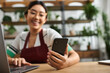 © LIGHTFIELD STUDIOS - A young Asian woman in an apron uses her phone while working in her plant store.