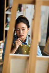 Wall Mural - A young Asian artist, wearing an apron, thoughtfully paints in her workshop.