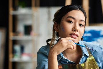 Wall Mural - A young Asian artist in an apron contemplates her next stroke, paintbrush in hand, in her art studio.