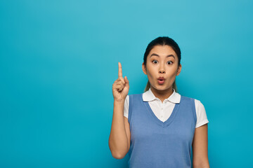 A young Asian woman in smart casual attire looks surprised with a raised finger, appearing to have just had a brilliant idea.