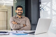 © Liubomir - Portrait of young arab businessman, man smiling and looking at camera while sitting inside office, boss in business suit at workplace using laptop