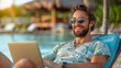 © Pinklife - A cheerful man wearing sunglasses and a vibrant shirt using a laptop while reclining at a poolside lounge