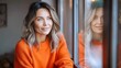 © Pinklife - Light-hearted and gracious woman smiles by the window draped in an orange sweater, exuding warmth