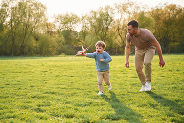 Wall Mural - Toy plane in hand, running. Happy father with son are having fun on the field at summertime