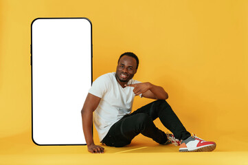 Wall Mural - Sitting near to the large smartphone with copy space. Black man is in the studio against yellow background