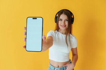 Wall Mural - Headphones on head, holding smartphone with copy space. Young woman is against yellow background