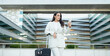 © Prostock-studio - A young woman in a white blazer and white pants stands in front of a modern building, holding a suitcase and looking at her smartphone. The woman is preparing for a business trip