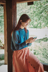 Wall Mural - Teenage girl using smartphone in a rustic wooden gazebo, absorbed in technology amidst nature. Casual lifestyle portrait with a focus on digital connection. Serenity and digital age concept.