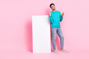 Poster - Full body photo of handsome young guy beaming smile hold gadget finger point banner wear trendy blue look isolated on pink color background