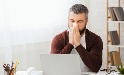 Aufkleber - Sick man sitting in front of a laptop is blowing his nose into a tissue. He appears to be dealing with a cold or allergies while using the computer.