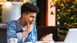 © Prostock-studio - A concentrated African American young guy is sitting at a table with a laptop in front of him, focused on the screen. He appears to be working or studying intently.