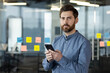 © Tetiana - Portrait of a serious young man standing in the office in a blue shirt, holding a phone in his hand and looking confidently into the camera