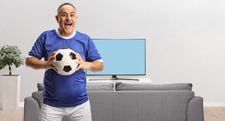 Wall Mural - Cheerful mature man in a football jersey holding a ball at home