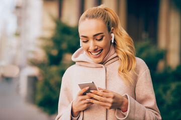 A young woman with blonde hair and a pink hoodie smiles while looking at her smartphone in an outdoor setting. She is wearing earbuds and appears to be enjoying her time on her phone.