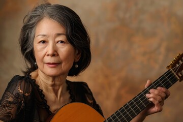 Wall Mural - Portrait of a tender asian woman in her 50s playing the guitar while standing against pastel brown background