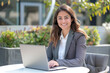 © pham - A woman in a suit sits at a cafe table, using her laptop with a bright smile on her face