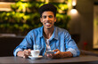 © Prostock-studio - A young black guy is seated at a table, where he is observed with a cup of coffee in front of him. He appears to be enjoying a moment of relaxation while sipping his beverage.