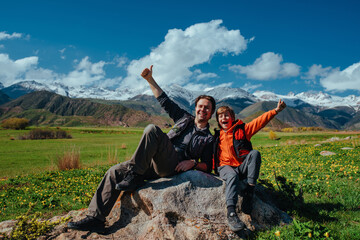 Canvas Print - Happy father and son hikers sitting on rock in the mountains and showing thumbs up