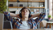 © Natali - A relaxed young man in headphones sits in the office at a table in front of a laptop and rests with his hands behind his head, listening to music