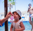 © Viga - A child wearing a blue and white bucket hat with yellow polka dots, a sleeveless ruffled white top, smiling warmly with a curious expression outdoors on a sunny day by the pool. Blurred background
