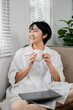© Satori Studio - Young woman in white shirt holding coffee cup, sitting on a couch with a laptop, smiling and enjoying a peaceful morning at home.
