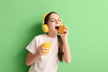 Wall Mural - Little girl in headphones with glass of juice eating orange on green background