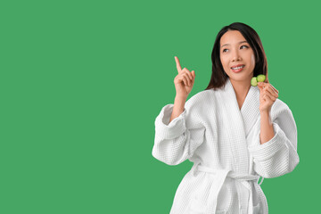 Wall Mural - Young Asian woman with cucumber slices pointing at something on green background