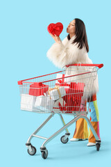 Poster - Beautiful young stylish Asian woman with shopping cart and gift boxes blowing kiss on blue background