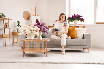 Wall Mural - Young woman sitting on sofa and using mobile phone near table with vase of lilacs in living room