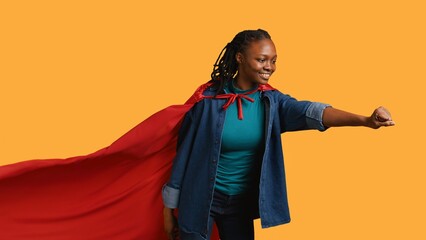 Happy girl portraying superhero flying with cape fluttering in wind, isolated over studio background. Upbeat heroic african american woman posing as hero in costume, camera B