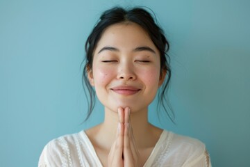 Wall Mural - Portrait of a happy asian woman in her 20s joining palms in a gesture of gratitude in front of soft blue background