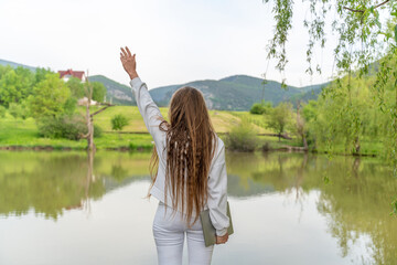 Wall Mural - A woman with long hair is standing by a lake, holding her hand up in the air. The scene is peaceful and serene, with the woman's pose and the calm water creating a sense of tranquility.