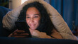 © Yuliia - Happy woman gadget addict social media addicted Hispanic smiling girl Latino Indian lady Arabian female suffering insomnia at night home relaxing under blanket cover with duvet browsing mobile phone