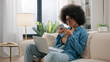 © Yuliia - African American woman confused puzzled pensive ethnic girl with laptop on couch browsing mobile phone thinking problem solution choose food delivery break in distant work scrolling smartphone at home