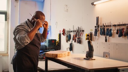 Carpenter inspecting wood in assembling shop, putting safety glasses on and picking tool from rack. Woodworking specialist in studio picking lumber block and carving gear for it, camera B