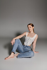 A young woman sits against a gray backdrop, wearing a white corset and blue jeans.