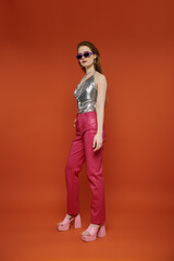 Wall Mural - A young woman in a silver top and pink pants poses against a vibrant orange backdrop.