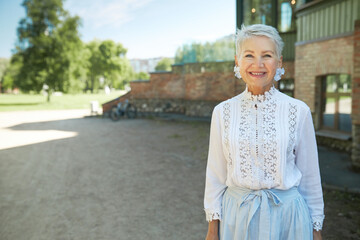 Wall Mural - Outdoor portrait with copy space for advertising content of retired stylish caucasian real estate agent posing against brick building with smile, wearing elegant laced blouse and skirt