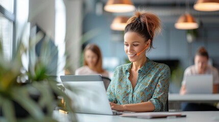 Wall Mural - Woman in a brightly lit modern office wearing a headset and working on a laptop with colleagues in the background to indicate a collaborative business environment