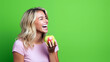© David Giraud - Belle femme blonde, heureuse et riant, mangeant une pomme sur un fond vert, portant un t-shirt rose.