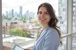 © pham - A smiling businesswoman in a blue blazer uses a tablet while looking toward the camera in a modern office setting. The city skyline can be seen through the window behind her