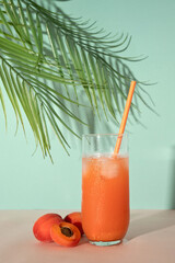 Wall Mural - Healthy refreshing summer cocktail drink with apricot fruit juice, ice and drinking straw on pastel peachy pink and turquoise blue background with soft sunlight shadow and palm tree leaves