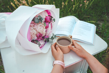 Canvas Print - A bouquet of peonies, an open book and a cup of tea in hands, a pleasant time in nature