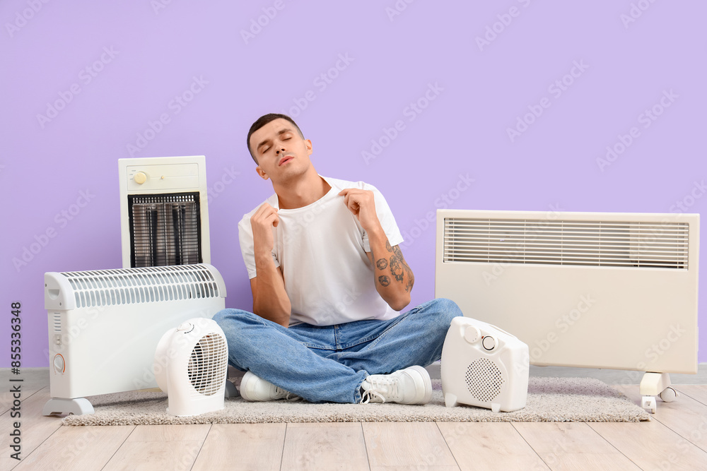 Young man with electric heaters sitting near lilac wall