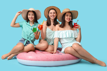 Wall Mural - Beautiful young women in wicker hats with glasses of cocktail sitting on inflatable mattress against blue background. Summer party