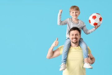 Wall Mural - Cute little boy sitting on father's shoulders with soccer ball and showing thumb-up on blue background