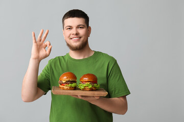 Wall Mural - Young man holding board with tasty burgers and showing OK on grey background